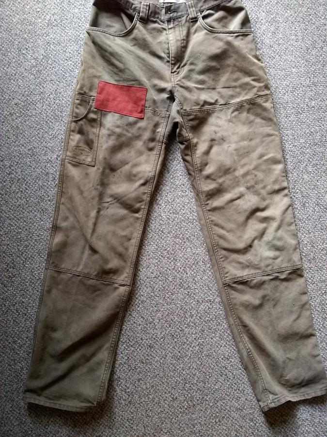 brown/gray work pants with upper thigh patched with reddish fabric, lying on gray rug