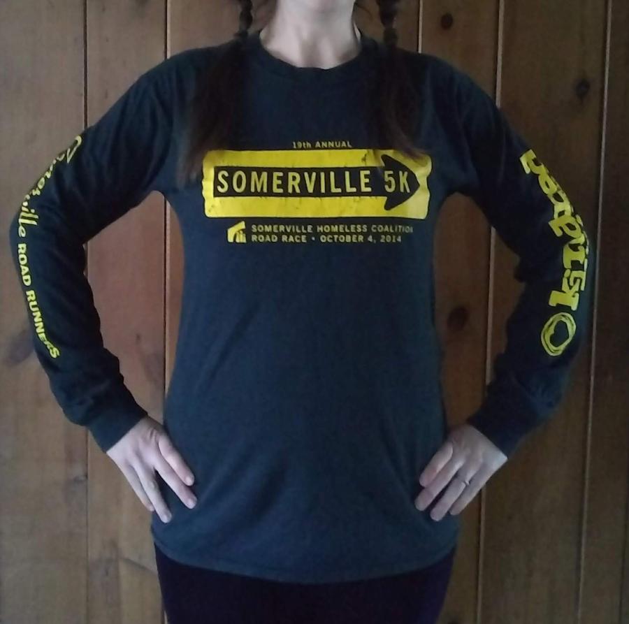 front view of woman wearing a straight, slim-fitting black long-sleeve t-shirt screen printed with the Somerville 5K logo