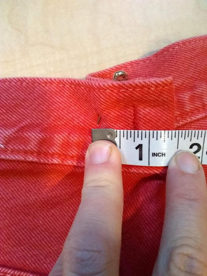 fingers holding down measuring tape over back waist/hip of red jeans, tape measures about 1.5 inches of excess fabric cinched in by safety pin