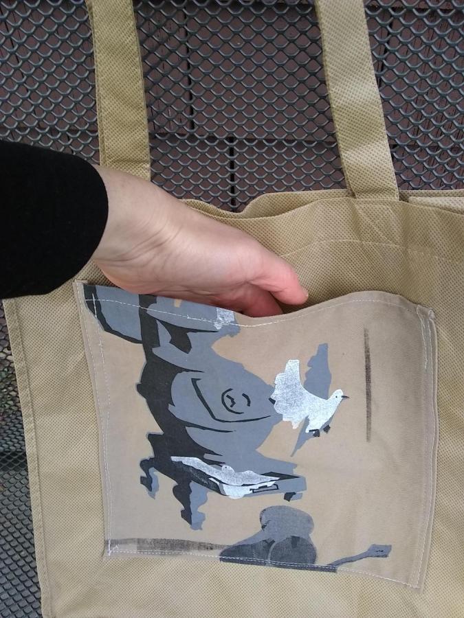 Beige tote bag with front hand indicating that there is a pocket on the front. Pocket has a silk screen of a woodstove and birds on it.