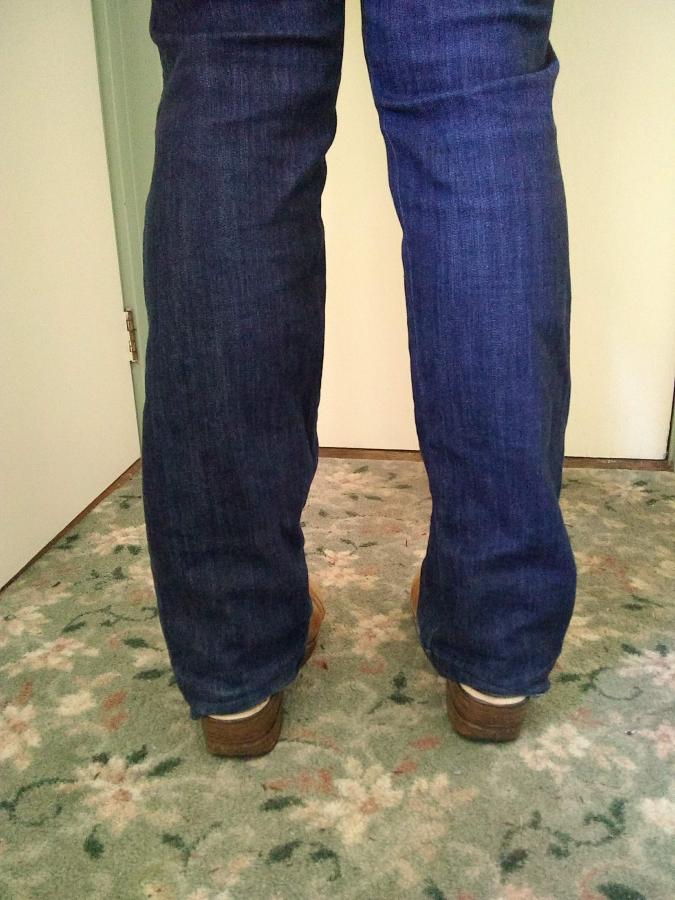 back view of woman's feet and ankles in too long dark blue jeans with wear at the hems