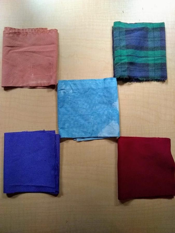 Five folded strips of fabric in quincunx pattern (i.e. like five dots on a die) on wooden background: terra cotta, black watch, blue tie dye, purple, red